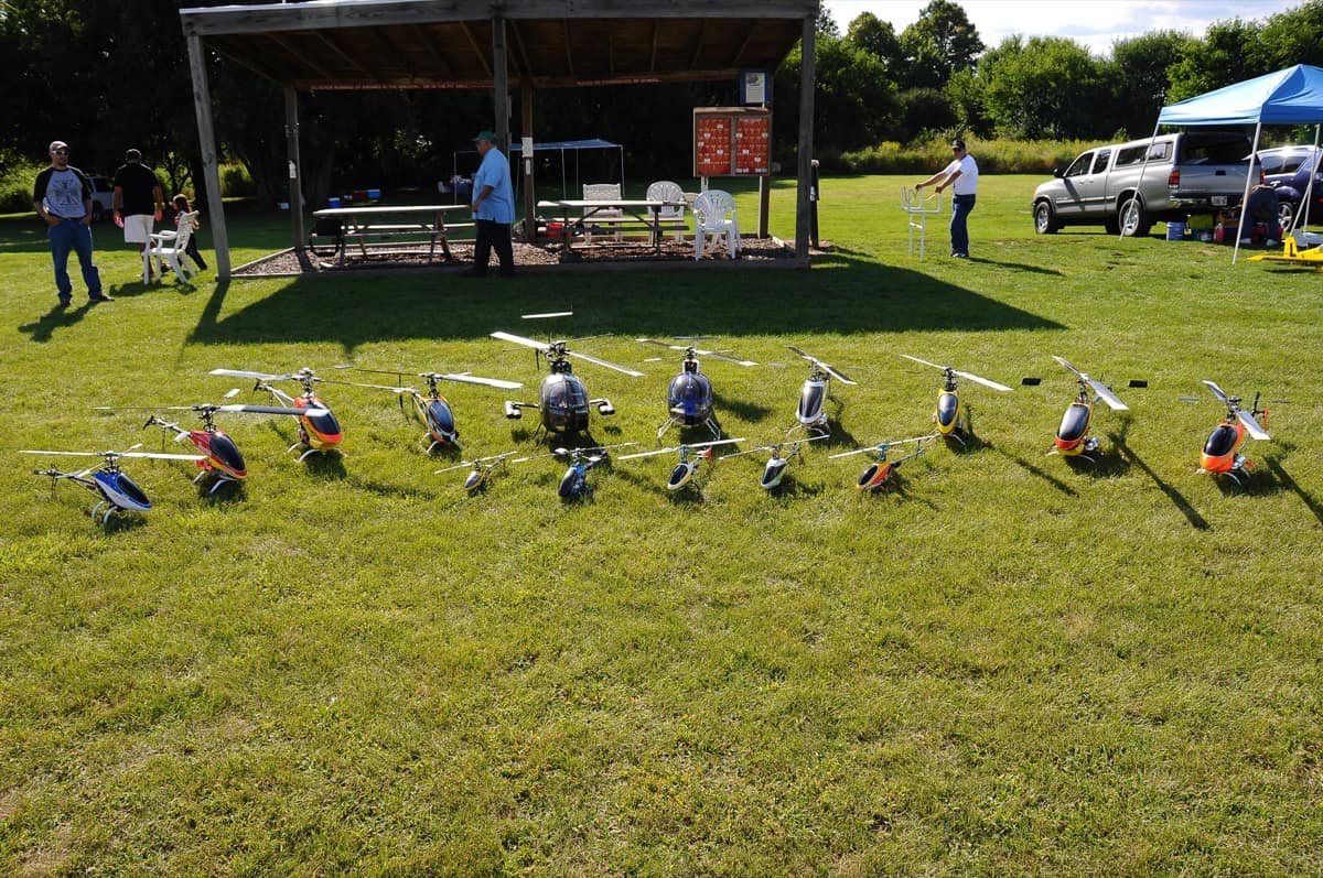 A lineup of RC helicopters at the field, with members and the club pavilion in the background