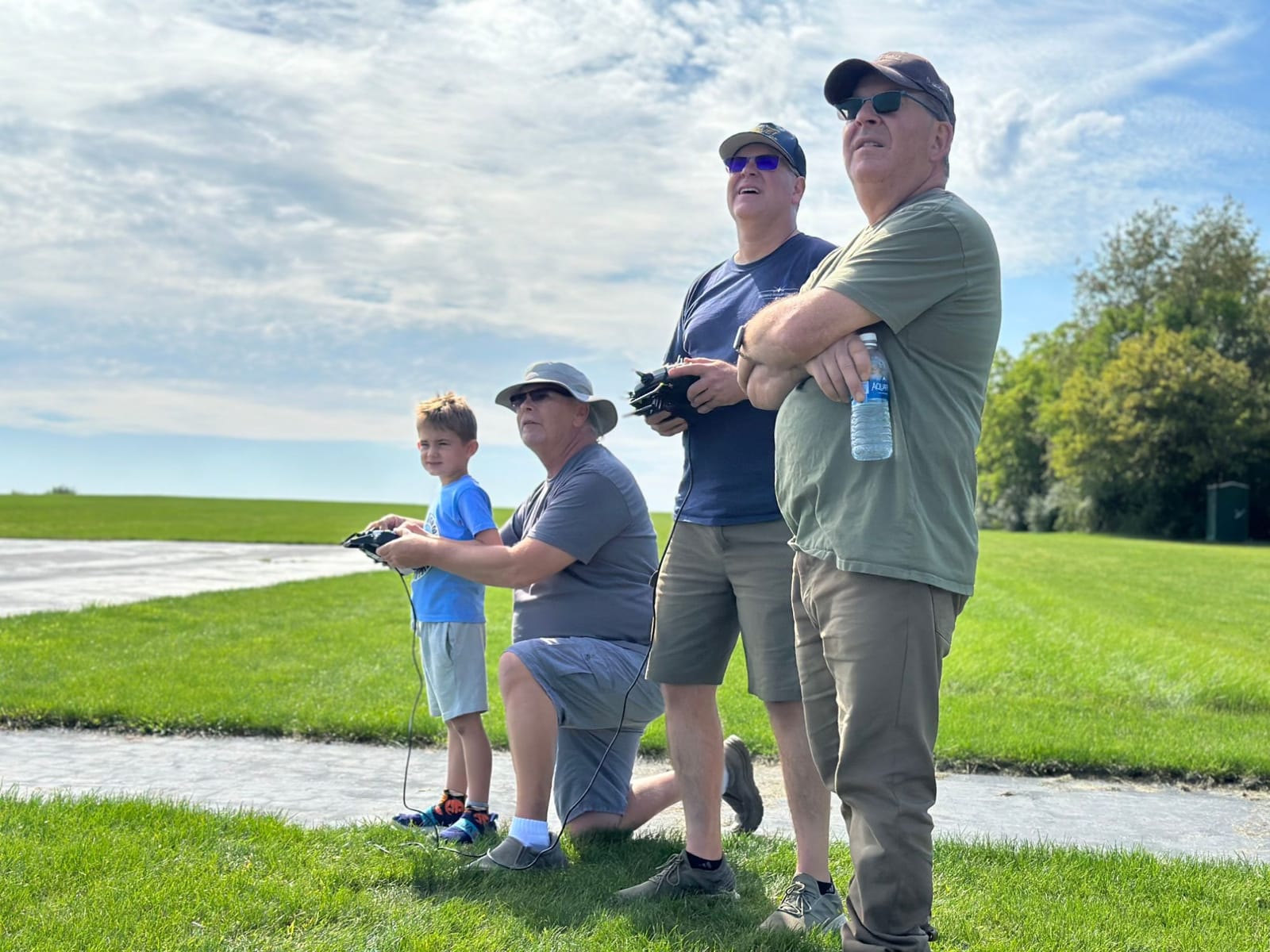 A young pilot holds a transmitter on a buddy-box cable while one instructor kneels beside him and another flies alongside, all watching the plane