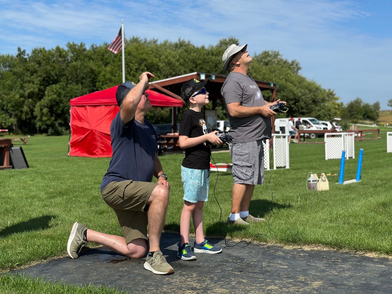 A boy flying on the buddy box looks up at his plane, with an instructor kneeling next to him and a second instructor holding the linked transmitter